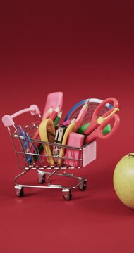 Vertical video of shopping trolley with school materials, apple and copy space on red background