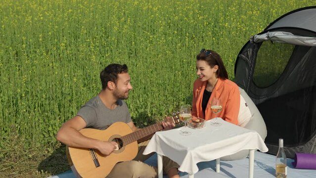 Romantic couple camping in countryside sitting by tent with drinks. Man Playing Music on Acoustic Guitar And Woman Drinking White Wine - shot in slow motion in Blooming Canola Field