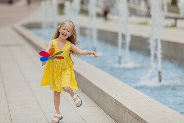 Happy little girl running in the park near the city fountain playing with paper windmill. Summer concept. happy childhood