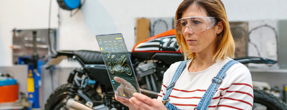 Concentrated female mechanic with security glasses touching transparent digital tablet with diagnosis app to review motorcycle on factory - Powered by Adobe