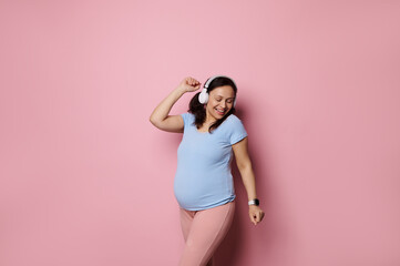 Happy beautiful cheery pregnant woman in blue t-shirt, dancing, listening to music on headphones,...