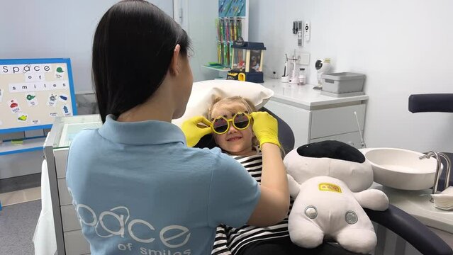 pediatric dentistry doctor puts yellow sunglasses on girl a girl in striped blouse with soft toy sits in dental chair in background board 