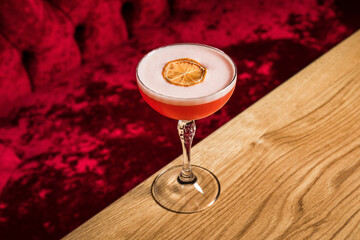 A pink sour alcoholic cocktail with foam garnished with dried lemon wheel served in a coupe glass on a wooden table of restaurant or bar - 608138467