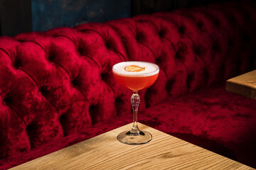 A pink sour alcoholic cocktail with foam garnished with dried lemon wheel served in a coupe glass on a wooden table of restaurant or bar - obrazy, fototapety, plakaty