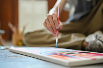 A male artist paints watercolors on a canvas while sitting on the floor in his studio.