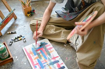 A male artist painting watercolors on a canvas while sitting on the floor in his studio.