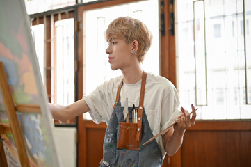 A creative Asian queer artist focuses on painting watercolors on a canvas easel in his gallery.