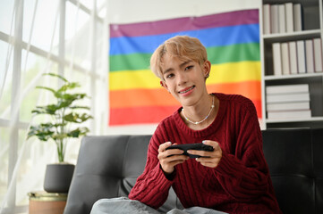 A smiling young Asian gay man sits on a sofa with his smartphone in his hands