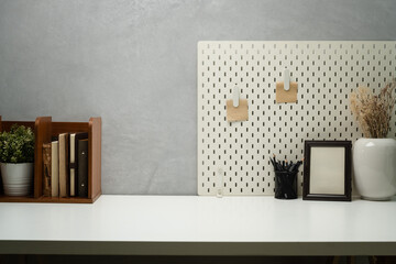 Stylish workplace with books, picture frame, pencil holder and pegboard on wall