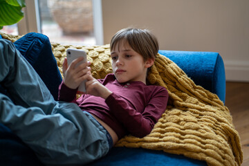 Interested little schoolboy using smartphone. Curious child holding cellphone in hands lying on couch downloads application at home alone. Relaxed boy playing mobile device game, watching cartoons