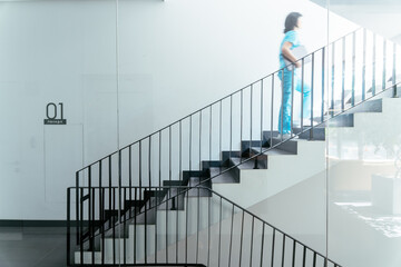 Woman wearing blue medical uniform going up stairs in modern clinic, motion blur. Medicine, profession and healthcare concept.