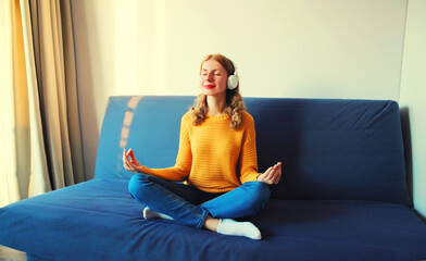 Happy relaxed young woman listening to music with wireless headphones meditating with closed eyes sitting in the lotus position on the couch at home