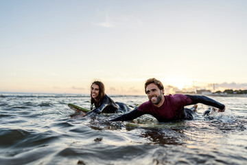 Happy couple of surfers swimming with boards in water.