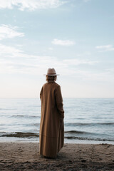 Lonely senior woman alone standing on seashore and looking thoughtfully into horizon. Back view of stylish elderly beautiful lady wearing hat, brown coat, mood of loneliness in nature