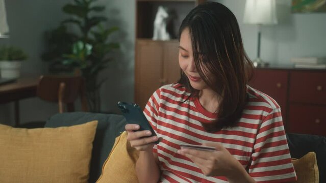 Attractive young asian woman shopping online using smartphone holding credit card purchase via e-banking. Beautiful girl making online payment through mobile e-commerce platform in cozy living room.