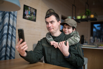Military man in olive uniform making selfie with his little son and smiling at home