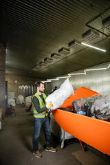 Side view of bearded male worker in glove and fluorescent vest putting sack with trash in container in blurred waste disposal station, garbage sorting and recycling concept