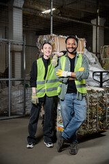 Obraz na płótnie Canvas Positive young sorter in protective vest and gloves looking at camera while standing near indian colleague and blurred waste paper in waste disposal station, garbage sorting and recycling concept