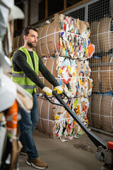 Bearded male worker in protective vest and gloves using hand pallet truck while standing near waste...