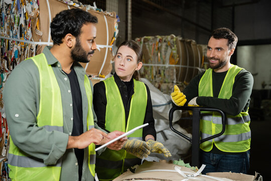 Smiling worker in reflective vest and gloves talking to multiethnic colleagues with digital tablet and standing near waste paper on hand pallet truck in garbage sorting center, waste recycling