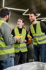 Positive workers in high visibility vests and gloves talking to indian colleague while standing near sack and working together in waste disposal station, recycling concept