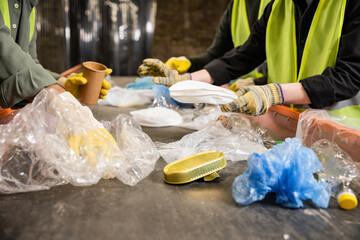 Cropped view of workers in high visibility vests and gloves separating different plastic and paper trash on conveyor while working in waste disposal station, recycling concept