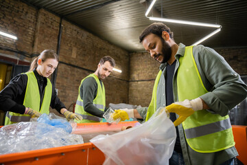 Indian worker in protective vest and gloves putting plastic trash in bag while working near blurred colleagues and conveyor in waste disposal station, garbage sorting and recycling concept