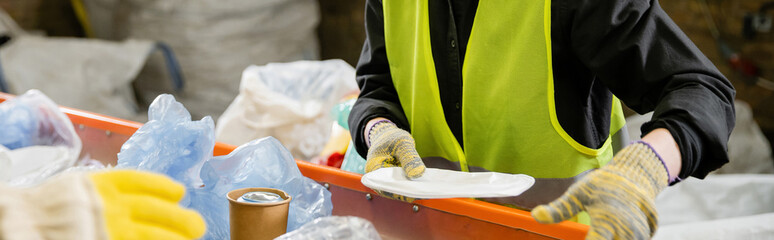 Cropped view of sorter in protective vest and gloves taking garbage from conveyor while working in...