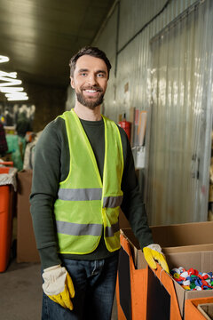cheerful sorter in high visibility vest and protective gloves looking at camera while standing near plastic caps in carton boxes in waste disposal station, garbage sorting and recycling concept
