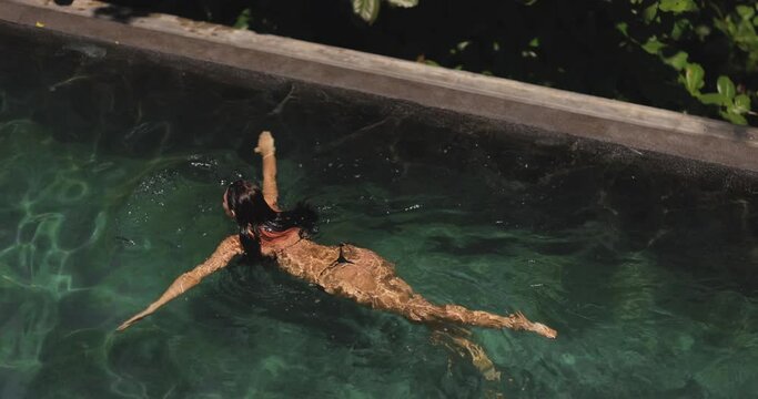 Aerial drone view of a slim young adult sexy woman in black bikini swimming in a luxury tropical resort pool. Slow motion 4k footage