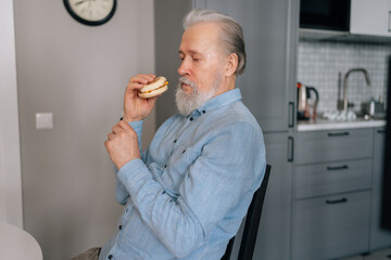 Side view of handsome gray-haired mature adult male eating unhealthy food enjoying dinner at home. Hungry bearded senior man eating delicious burger sitting at table in kitchen room.
