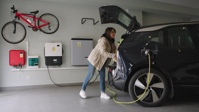 Woman parks in the garage and plugs a charger into socket of the electric car. Renewable houses, Energy Efficient Domestic Life. Horizontal Video 4k.