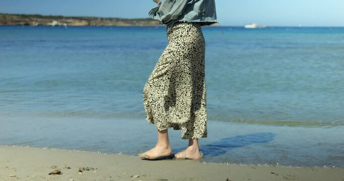 Anonymous fashionable woman make slow graceful step on sandy beach in promenade. Student girl barefoot walk on sea coastline in light breeze wind on summer day.