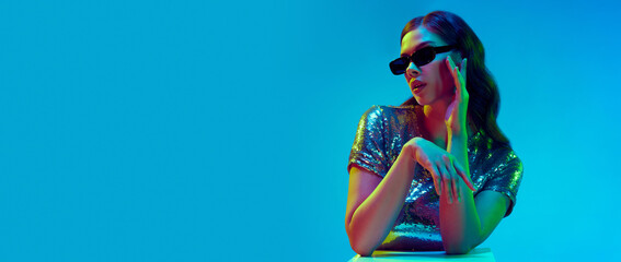 Banner with attractive fashion model, dreaming girl wearing trendy clothes, silver top and black sunglasses over blue background in neon light. Concept of beauty, fashion week - obrazy, fototapety, plakaty