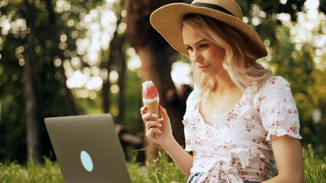 Young Blonde Woman In Straw Hat Working Or Studying On Laptop While Sitting On Grass In Park And Eating Ice Cream, Outdoor Freelancer, Distance Learning