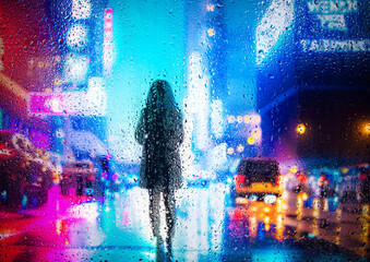 View through a glass window with raindrops on a blurred silhouette of a people under an umbrella walking on autumn rain , night street scene. focus on raindrops