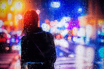 View through glass window with rain drops on blurred reflection silhouette of a girl on a city street after rain and colorful neon bokeh city lights, night street scene. Focus on raindrops on glass