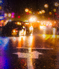 View through the glass covered with raindrops on the streets of a big city in the autumn rain. lights of car headlights and windows of houses reflected in puddles on the road. Focus on drops on glass