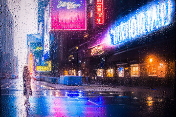 View through glass window with rain drops on blurred reflection silhouettesof a girl in walking on a rain under umbrellas and bokeh city lights, night street scene. Focus on raindrops on glass	