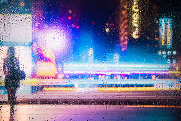 View through glass window with rain drops on blurred  silhouette of a girl on a city street after rain and colorful neon bokeh city lights, night street scene. Focus on raindrops on glass