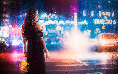 View through glass window with rain drops on blurred  silhouette of a girl on a city street after rain and colorful neon bokeh city lights, night street scene. Focus on raindrops on glass