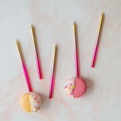 Party candles in pink and gold, with two colorful macarons, against a pink marble background
