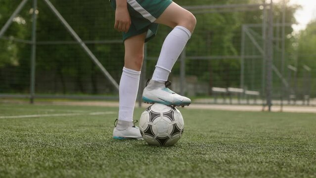 The boy dressed in football uniform is practicing on a mini football stadium. A close-up shot of the legs of the young footballer, with one foot is on the ball