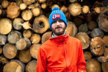 Man in a blue beanie and a red jacket poses against a background of cut and stacked wooden logs forming a pattern