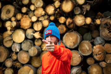 Man in a blue beanie and a red jacket poses against a background of cut and stacked wooden logs forming a pattern