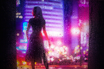 View through glass window with rain drops on blurred  silhouette of a girl on a city street after...