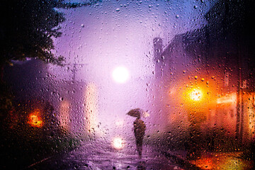 View through a glass window with raindrops on a blurred silhouette of a girl with umbrella walking...