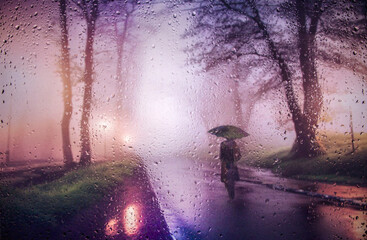 View through a glass window with raindrops on the silhouette of a man walking under an umbrella down the street in the morning mist in the rain Colorful lights of street lamps Focus on the raindrops