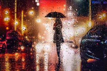 View through a glass window with raindrops on a blurred silhouette of a girl with umbrella walking on autumn rain , night street scene. focus on raindrops