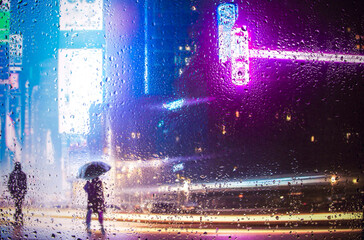 View through glass window with rain drops on blurred reflection silhouette of a girl on a city street after rain and colorful neon bokeh city lights, night 
